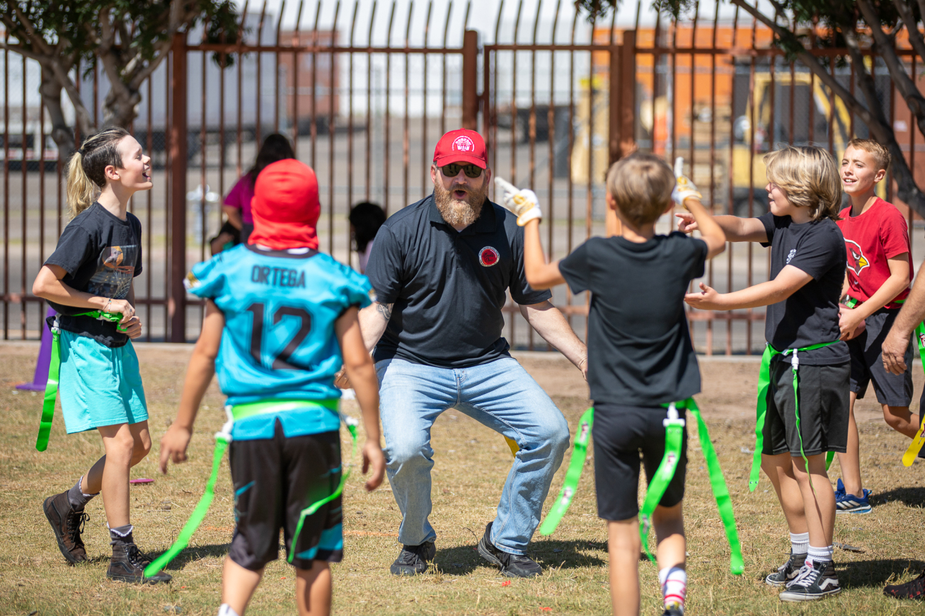 A dad in a red baseball hat and black shirt and jeans stands on the field with kids around him during the flag football game