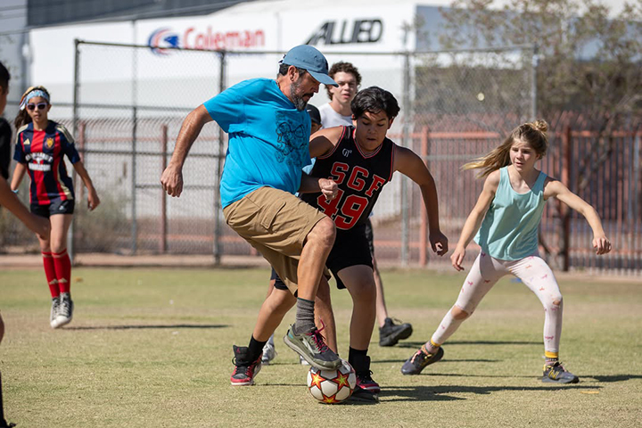 A man in a blue shirt and a boy in a jersey try to get the soccer ball