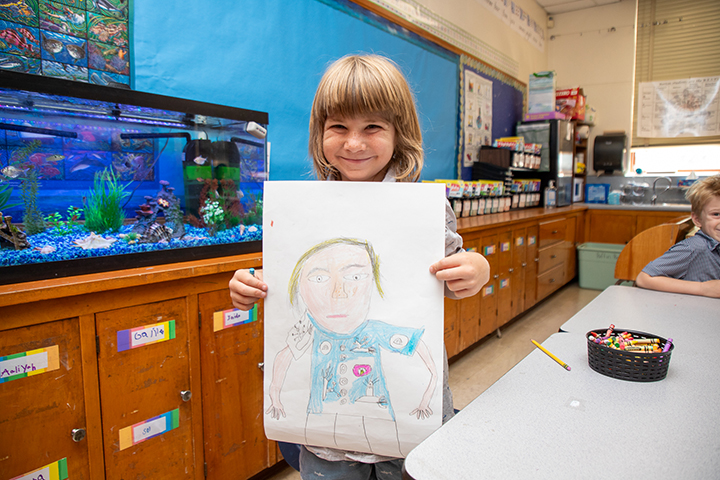 A little boy holds up his self-portrait next to an aquarium