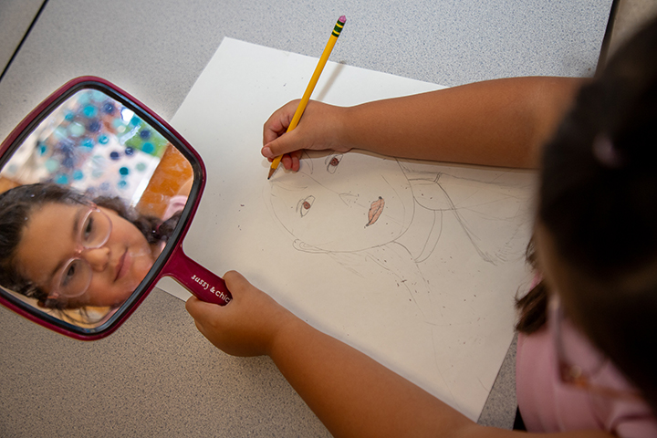 A girl holds up a mirror as she draws a self-portrait