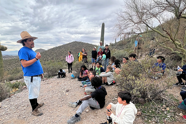 A man talks to a group of students who are sitting on the ground in the desert