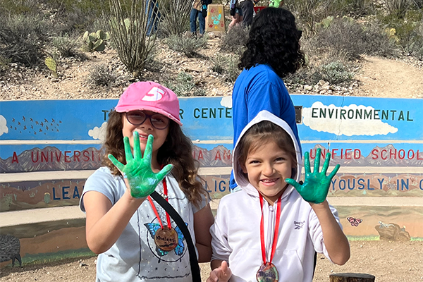 Two girls smile as they hold up their hands covered in green paint
